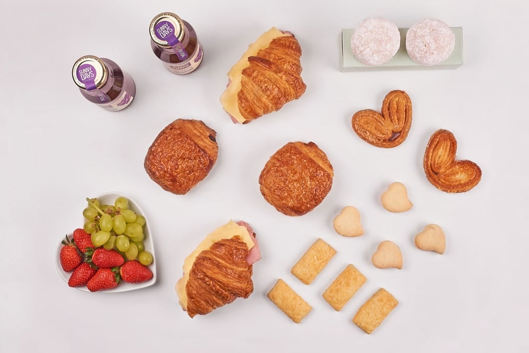 Box de café da manhã com croissants, pães, frutas e sucos.